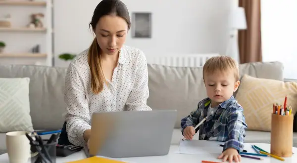 Kobieta pracuje przy laptopie, podczas gdy jej syn rysuje obok. To obrazek z życia, gdzie praca i rodzina łączą się, jak w przypadku art. 188 kodeksu pracy.