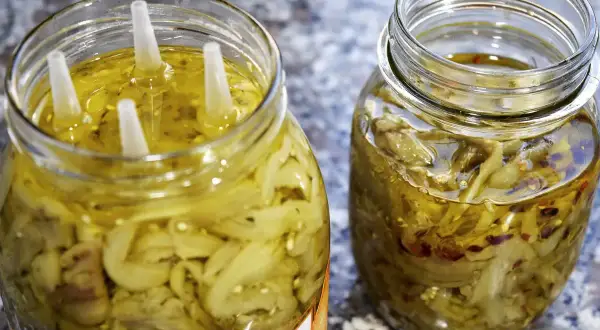 Due barattoli di melanzane a filetti sott'olio, uno con quattro tappi bianchi che emergono dall'olio dorato.
