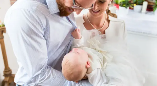 Szczęśliwi rodzice trzymają niemowlę w białej szatce. W tle kościół, gdzie odbywają się chrzciny dziecka. Przesądy związane z tym wydarzeniem są różne.
