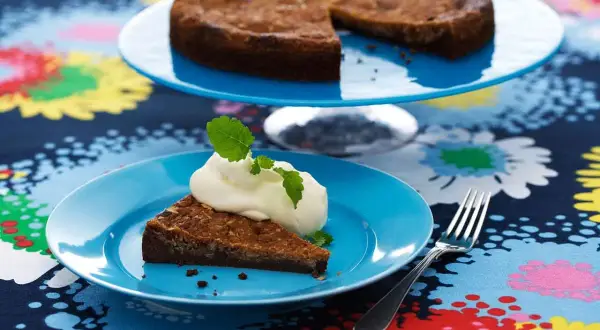 En bit kladdkaka med vispad grädde och mynta på en blå tallrik. Hela kladdkakan syns på en tårtfat i bakgrunden. Ett perfekt kladdkaka recept för fikat.