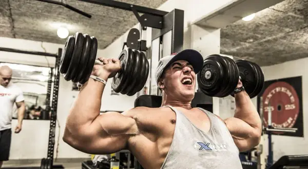 Man med keps och linne lyfter tunga hantlar i ett gym. Han pressar sig till max med varje repetition i sitt cluster set.