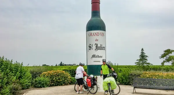 Zwei Radfahrer pausieren vor einer riesigen Weinflasche in Medoc, Frankreich. Weinberge erstrecken sich bis zum Horizont.