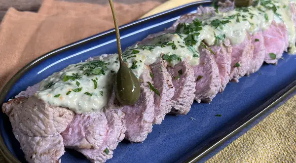 Tranches de vitello tonnato, nappées d'une sauce crémeuse et garnies de câpres et de persil, sur un plat bleu.