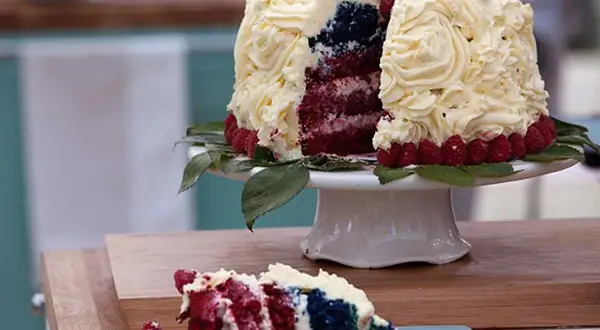 En skivad red velvet cake med vit frosting, hallon och blått inuti. En bit ligger på en blå tallrik.