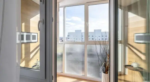 Zabudowany balkon z widokiem na budynki, ozdobiony wazonem z gałązkami.