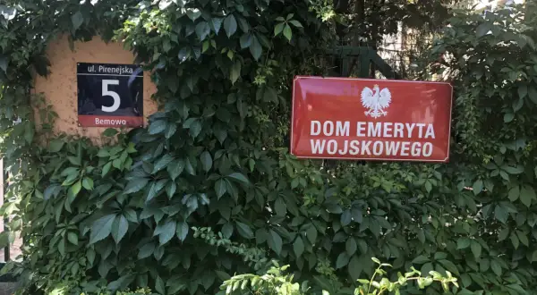 Czerwona tablica z godłem Polski i napisem "DOM EMERYTA WOJSKOWEGO" wśród zieleni. Obok numer domu 5.