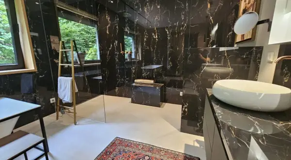 Luxuriöses Bad ohne Fenster mit schwarzem Marmor, freistehender Badewanne, Dusche und Waschbecken. Ein bunter Teppich liegt auf dem Boden.