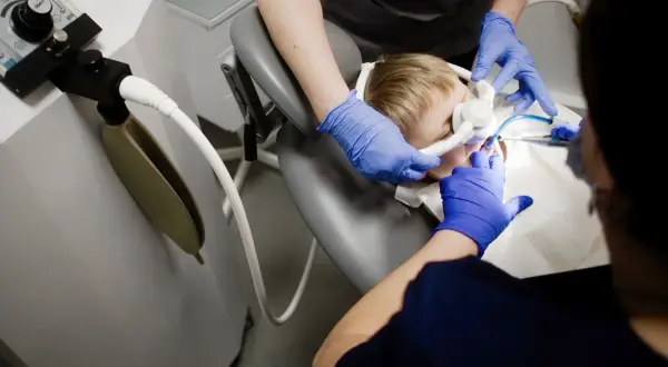 Chłopiec w masce do znieczulenia ogólnego u dentysty. Dwie osoby w niebieskich rękawiczkach wykonują zabieg.
