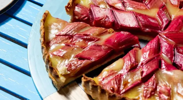Une tranche de tarte à la rhubarbe, garnie de fruits rouges et d'une crème dorée, repose sur une assiette bleue.
