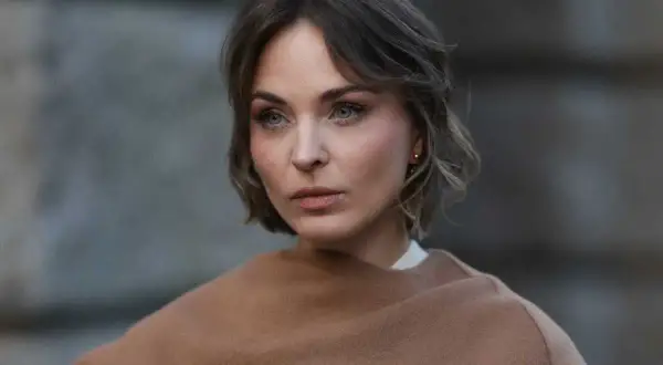 Eine Frau mit Kurzhaarfrisuren und einem leicht molligen Gesicht blickt nachdenklich in die Ferne, eingehüllt in einen braunen Schal.
