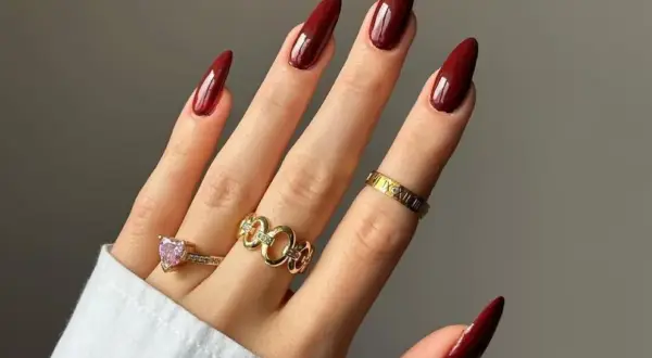 Main d'une femme aux ongles longs et pointus, vernis d'un bordeaux profond, parfaite tendance ongle automne. Bagues dorées et une bague cœur rose ornent ses doigts.