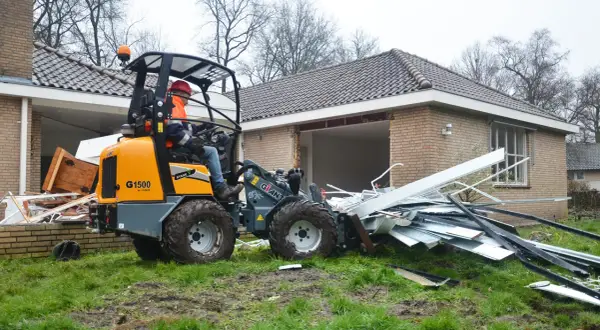 Pomarańczowa koparko-ładowarka z operatorem w kasku i kamizelce usuwa gruz z posesji.