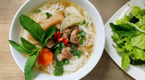 Aromaticzny makaron pho z kawałkami mięsa, warzywami i świeżymi ziołami. Obok talerz z sałatą i kiełkami fasoli.