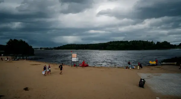 Plaża nad jeziorem Wysoka, z ciemnymi chmurami na niebie. Ludzie spacerują po piasku, niektórzy odpoczywają. W tle las i spokojna tafla wody.