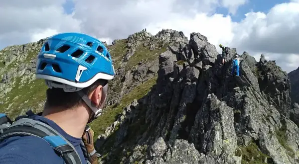 Alpinista w niebieskim kasku patrzy na grań Tatr, gdzie wspina się grupa ludzi.
