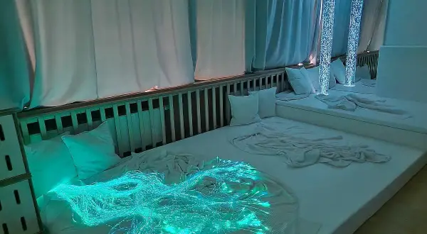 Ein beruhigender Snoezelen Raum mit weißen Matratzen, Kissen und faszinierenden Lichteffekten, darunter leuchtende Glasfasern.