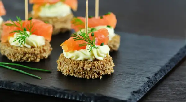 Eleganckie finger food: krążki ciemnego pieczywa z kremowym serkiem, łososiem i koperkiem, podane na łupkowej desce.