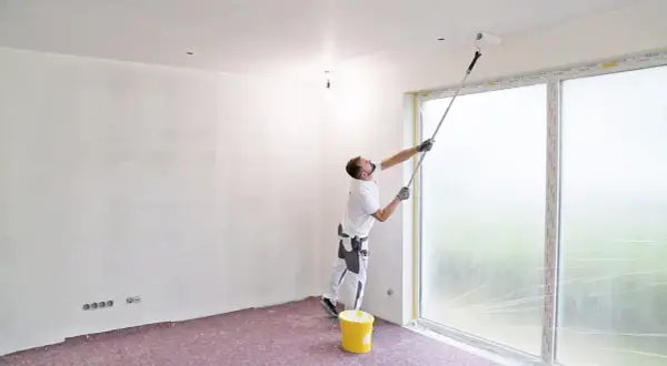 Maler streicht die Decke mit einer Teleskopstange. Ein gelber Farbeimer steht bereit.