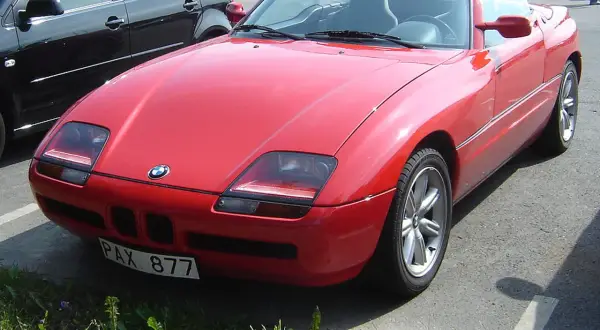 Czerwony kabriolet BMW Z1 z tablicą rejestracyjną PAX 877 zaparkowany obok czarnego samochodu.