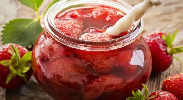 Ein Glas mit ganzen Erdbeeren und Sirup, bereit zum Erdbeeren einwecken. Frische Erdbeeren und Blätter liegen daneben.