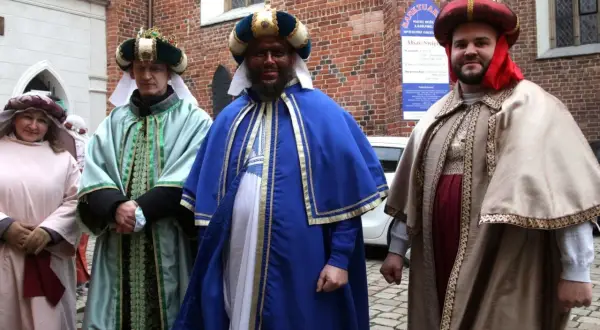 Trzej królowie w barwnych strojach i nakryciach głowy pozują do zdjęcia na tle ceglanej ściany.