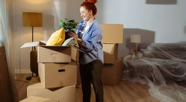 Donna con pianta in mano, circondata da scatoloni, in una casa in fase di trasloco. Potrebbe essere interessata alle agevolazioni prima casa senza residenza.