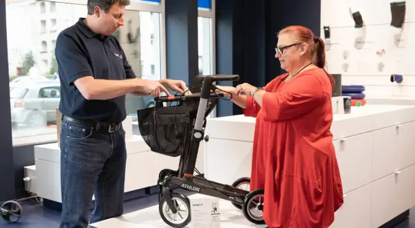 Mann erklärt einer Frau einen Rollator. Eine medizinische Hilfsmittel Liste ist im Hintergrund zu sehen.