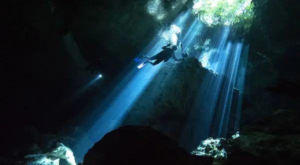 Buceadores exploran la asombrosa profundidad de un cenote, con rayos de luz solar atravesando la oscuridad.