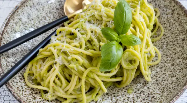 Cremige Avocado-Pasta mit frischem Basilikum und Parmesan auf einem rustikalen Teller, serviert mit schwarzem Besteck.