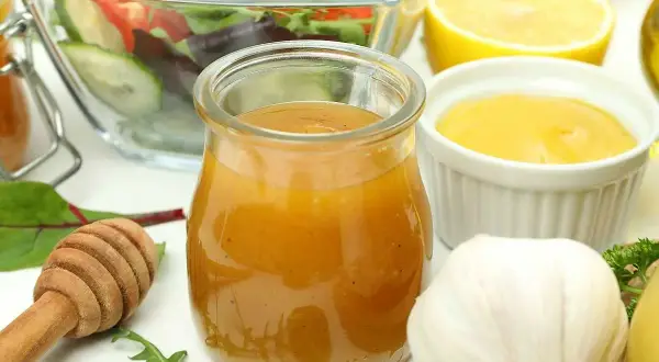 En burk med gyllene salladsdressing, bredvid en skål med sallad, citron, vitlök och honung. Ett perfekt salladsdressing recept!