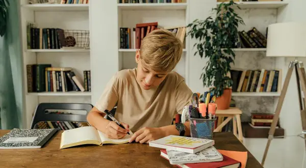 Un niño concentrado escribe en su cuaderno, rodeado de libros y útiles escolares, practicando dictados cortos para mejorar su ortografía.