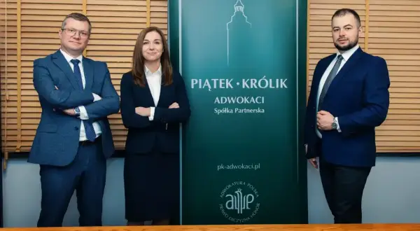 Trzy osoby w garniturach stoją przed zielonym banerem kancelarii "Piątek • Królik Adwokaci".