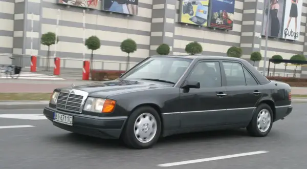 Ciemnoszary Mercedes W124 jedzie ulicą przed budynkiem z banerami.