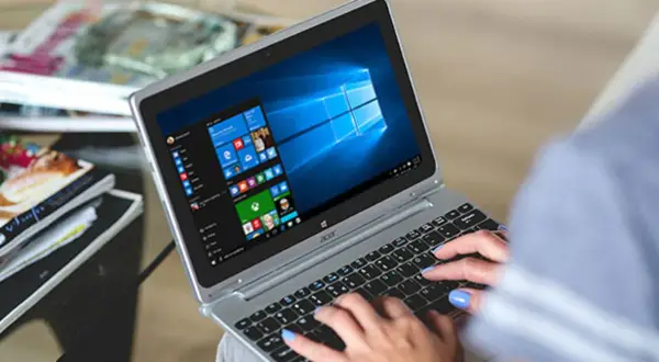 Kobieta pisze na laptopie z systemem Windows 10. W tle widać stosy czasopism, co sugeruje, że zadania w tle ochrony systemu są wykonywane podczas relaksu.