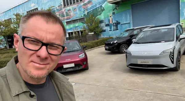 Mężczyzna w okularach pozuje do selfie na tle trzech samochodów marki AION.