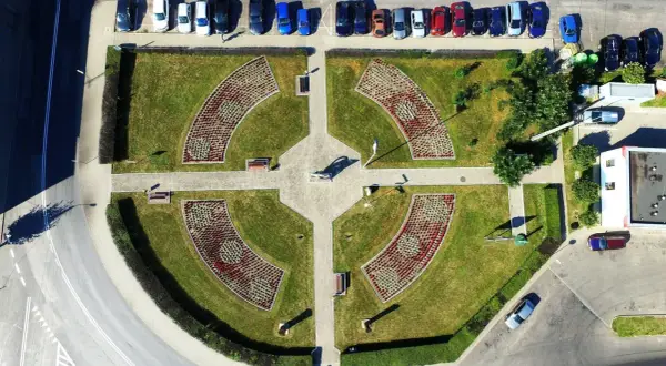 Piątek miasto środek Polski. Park miejski z klombami w kształcie łuków, alejkami i ławkami. W tle parking z samochodami.