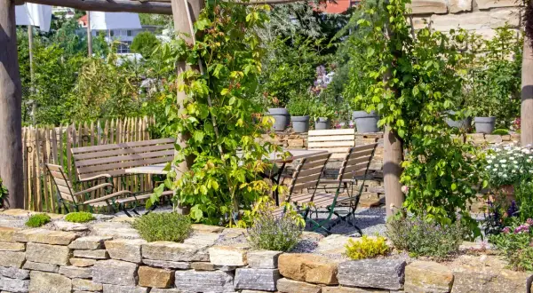 Un angolo di giardino con muretti a secco, un pergolato in legno, tavoli e sedie, e un'aiuola fiorita.