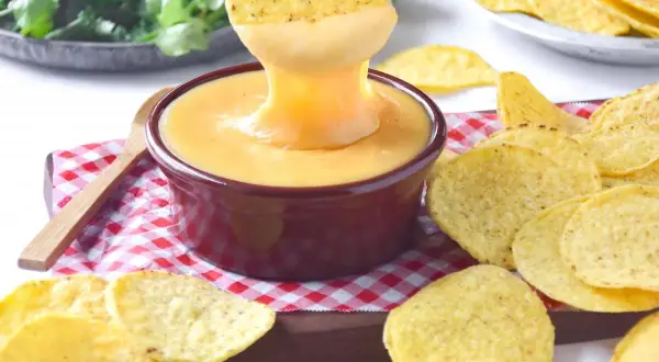 Un totopo se sumerge en una cremosa salsa cheddar, rodeado de más totopos y cilantro fresco.