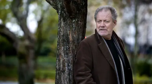 Harald Bovallius lutar sig mot ett träd i en park, klädd i brun rock och halsduk.