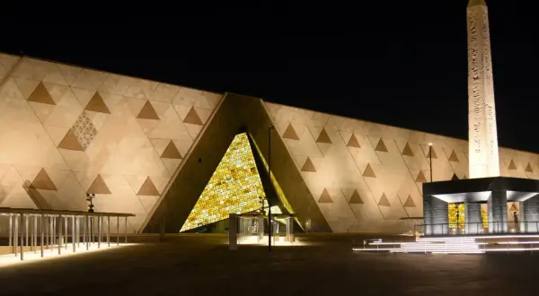 Nowe muzeum w Egipcie nocą. Monumentalna fasada z geometrycznymi wzorami i podświetlonym wejściem. Obok stoi starożytny obelisk.