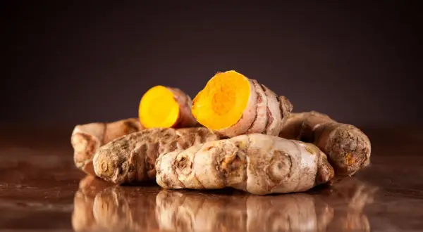 Ein Haufen frischer Kurkumawurzeln, eine davon aufgeschnitten, enthüllt ihr leuchtend orangefarbenes Inneres und die wertvollen Kurkuma Inhaltsstoffe.