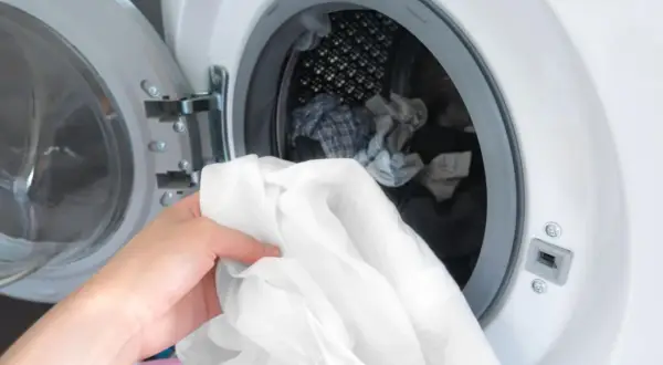 Eine Hand hält ein weißes Hemd, bereit, es in die Waschmaschine zu legen. Die Tür der Waschmaschine ist geöffnet und gibt den Blick auf weitere Wäsche frei.