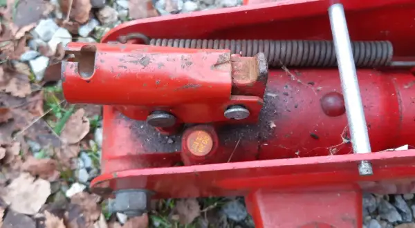 Detaljbild av en röd domkraft. En metallpinne sticker upp och en fjäder är synlig. Det ser ut som att det är dags att fylla olja i domkraften för att den ska fungera som den ska.