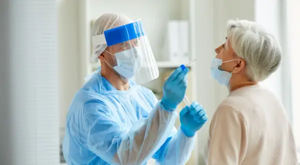 Arzt in Schutzkleidung nimmt bei älterer Frau einen Rachenabstrich. Testet man so auch auf MRSA, was ist das?