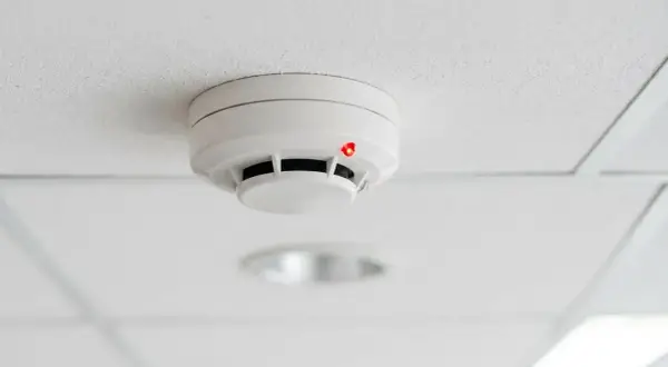 Détecteur de fumée blanc sur un plafond blanc, avec un voyant rouge allumé, suggérant un détecteur qui se déclenche tout seul.