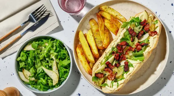 En härlig fransk mat-inspirerad måltid med en baguette fylld med krämig sås, ruccola, päron och knaprigt bacon, serverad med klyftpotatis och en fräsch sallad.