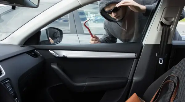 Un homme en capuche et lunettes de soleil tente un vol à la roulotte, outil rouge à la main, regardant à l'intérieur d'une voiture.
