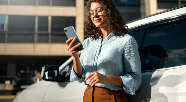 Kobieta w okularach przegląda dane w telefonie, opierając się o samochód. Może to być związane z systemem informacji kredytowej.