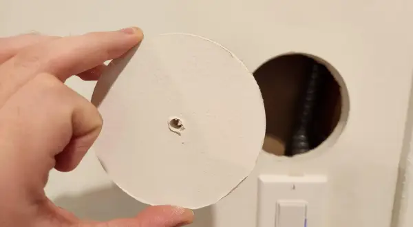 Un disco di cartongesso viene tenuto in mano vicino a un buco nel muro, con un interruttore della luce visibile sotto.
