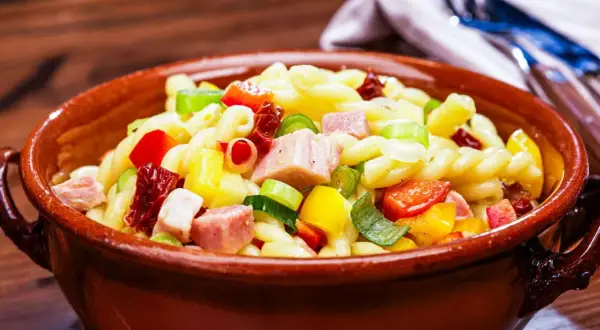 Leckerer nudelsalat ohne mayonnaise mit bunten Paprikawürfeln, Schinken und Frühlingszwiebeln in einer Tonschüssel.